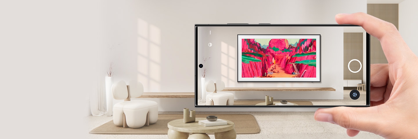 A hand holds up a smartphone in a living room. The smartphone screen shows how The Frame looks installed on the wall via AR.