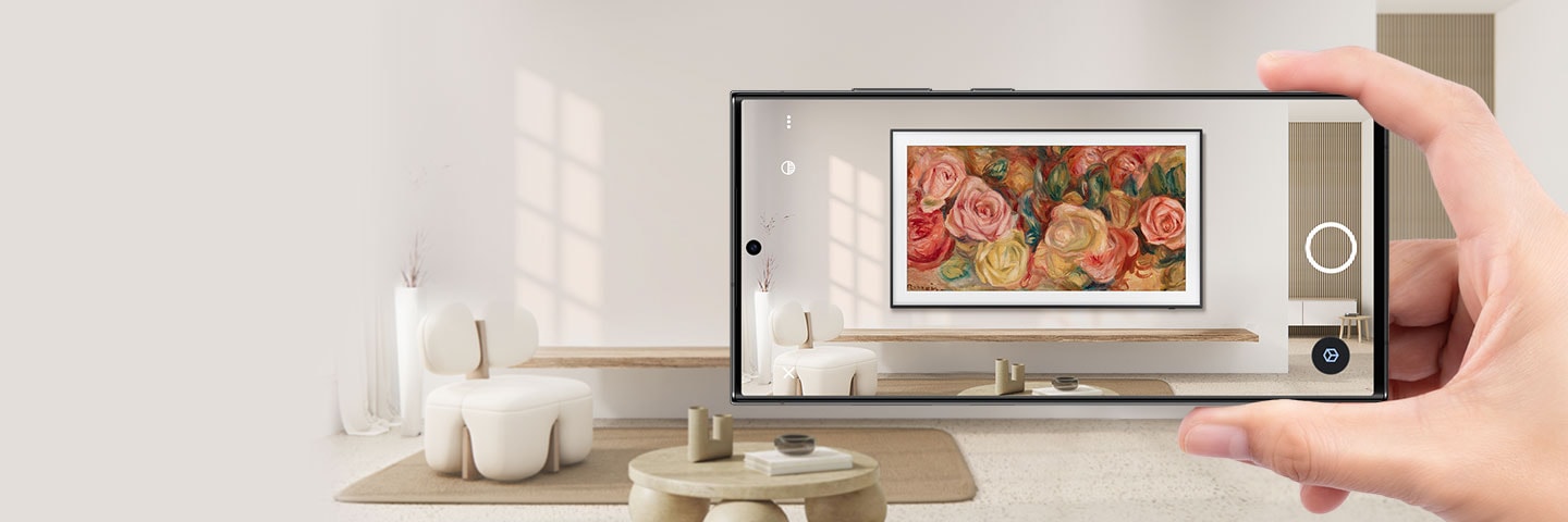 A hand holds up a smartphone in a living room. The smartphone screen shows how The Frame looks installed on the wall via AR.