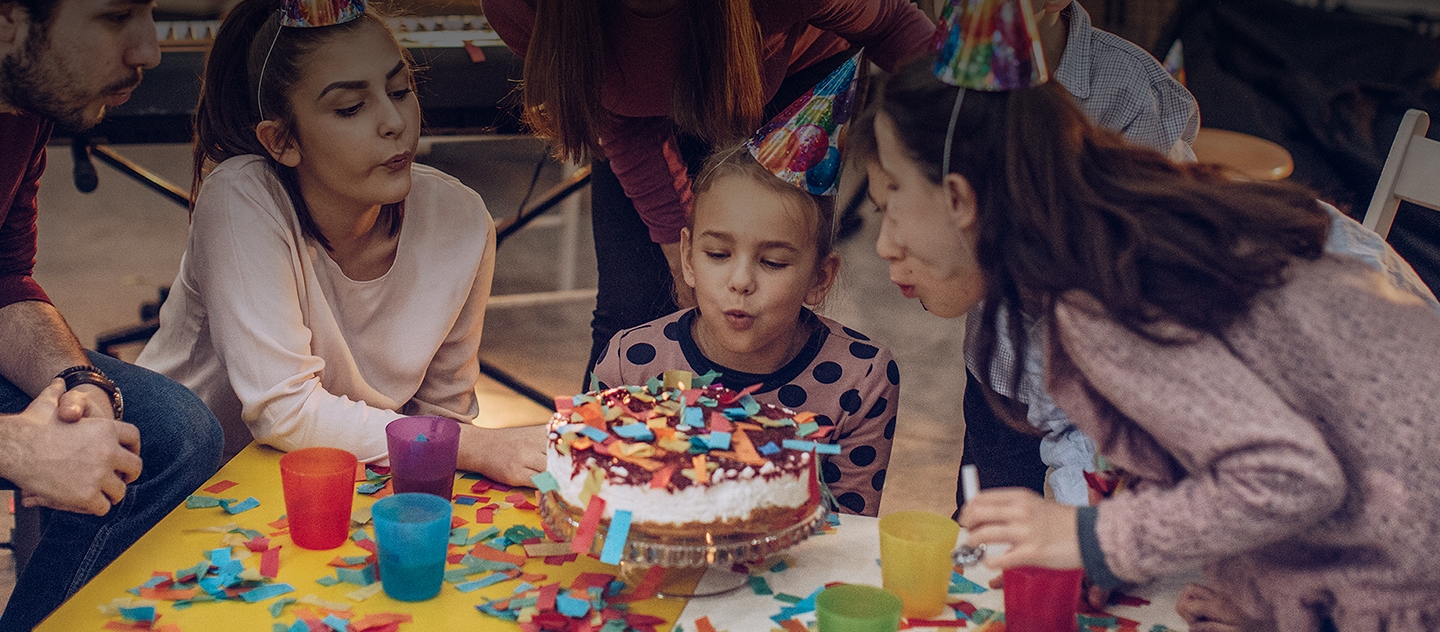 생일 파티에서 아이들과 어른들이 모두 생일 모자를 쓰고 있습니다. 색종이 조각이 흩날리는 가운데 모두 함께 중앙의 생일 케이크 촛불을 불어 끄고 있습니다.
