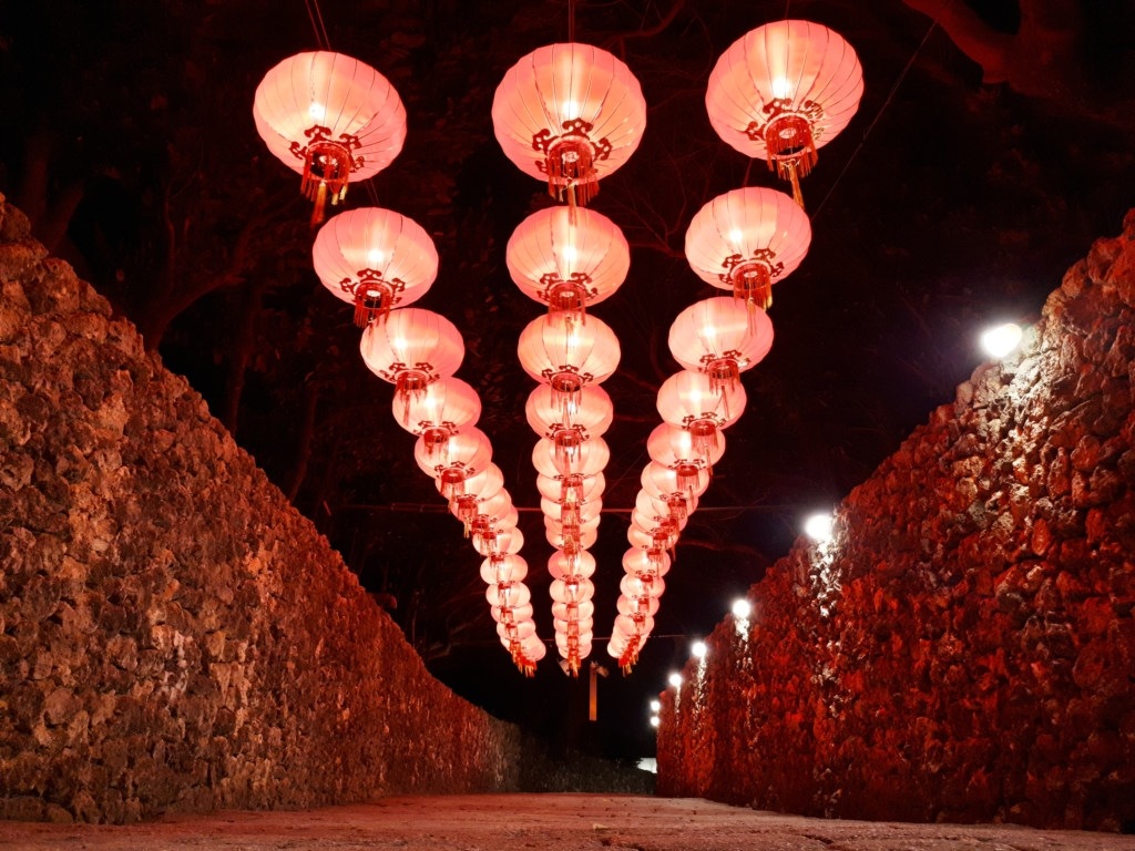 Streams of colorful traditional lanterns hang over a walkway flanked by stone walls.