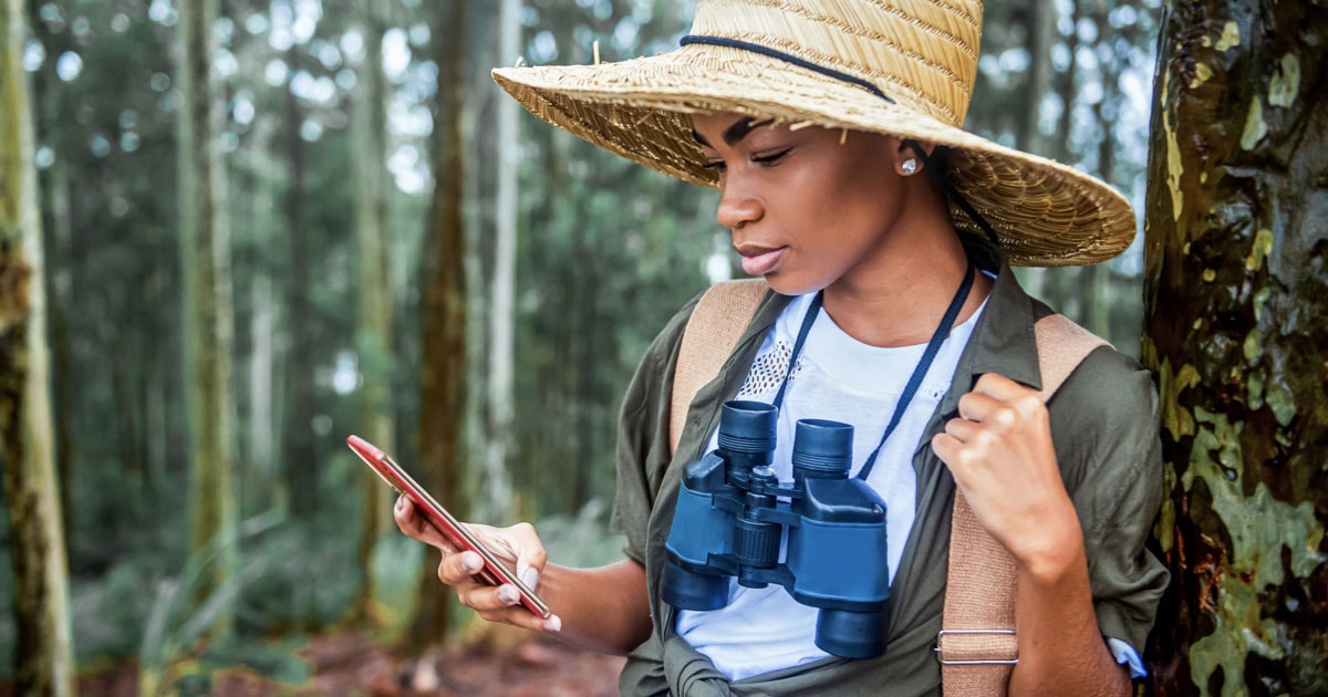 Eine Frau mit einem breitkrempigen Hut und einem Fernglas um den Hals schaut auf ihr Smartphone, während sie sich an einen Baum im Wald lehnt.