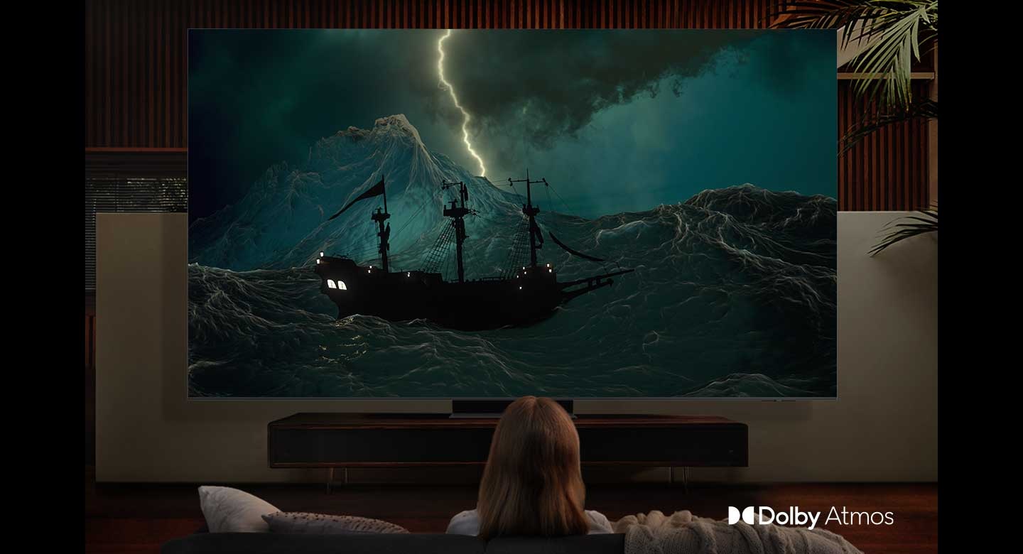 The tumultuous scene of a ship battling the sea amidst thunderstorms is showcased in a cinema. The very same scene is on display on Samsung's 98 inch Supersize TV within the living room, accompanied by cinematic sound facilitated by Dolby Atmos. The Dolby Atmos logo is prominently displayed in the lower right-hand corner of the 98 inch TV.