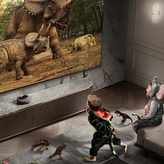 Duży telewizor Samsung wyświetlającym obraz dinozaurów w akcji, z dwiema osobami siedzącymi na sofie, oglądającymi film w realistycznej jakości