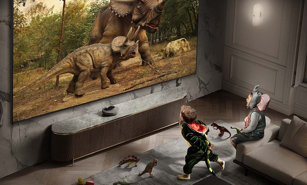 Duży telewizor Samsung wyświetlającym obraz dinozaurów w akcji, z dwiema osobami siedzącymi na sofie, oglądającymi film w realistycznej jakości